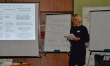 Жінка із світлим волоссям тримає записи у руках. На фоні дві дошки та екран.