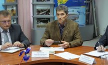 Чоловік у піджаку за круглим столом серед інших учасників, на фоні — інформаційні стенди.