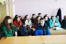 Студенти сидять в приміщенні за партами та слухають доповідача.