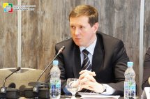 Чоловік у діловому костюмі сидить за столом та говорить у мікрофон.