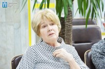 Жінка поважного віку з коротким світлим волоссям сидить у приміщенні.