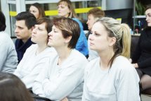 Три жінки у сірому одязі сидять у приміщенні. На задньому фоні видно інших людей.