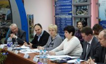 Люди сидять за столом у приміщенні. На задньому фоні - інформаційні стенди.