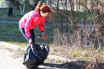 Жінка у рожевій куртці збирає сміття у чорний пакет у парку.