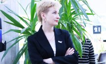 Жінка з короткою зачіскою у чорному одязі схрестивши руки перед собою перебуває у приміщенні.