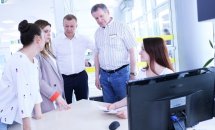 Жінка сидить за робочим місцем, біля неї стоять двоє чоловіків та дві жінки.