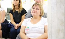 Жінка у білому одязі сидить у приміщенні, позаду сидить чоловік та жінка.