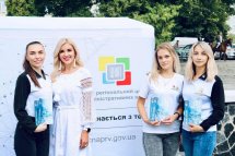 Група жінок у білих футболках позує на фоні банера на вулиці.