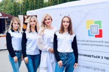 Група жінок у білих футболках позує на фоні банера на вулиці.