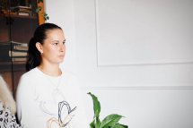 Дівчина у білому светрі сидить біля рослини, дивиться вбік.