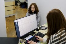 Дівчина сидить за комп’ютером, на екрані відкрита програма. Збоку сидить дівчинка