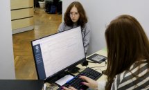 Дівчина сидить за комп’ютером, на екрані відкрита програма. Збоку сидить дівчинка