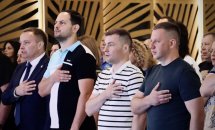 Група людей стоїть у приміщенні з дерев’яним інтер’єром, поклавши руки на серце.