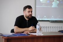 Чоловік у чорній футболці сидить за столом та говорить, на фоні екран із презентацією.