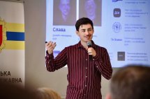 Чоловік у бордовій сорочці виступає перед аудиторією, на екрані за ним – презентація.