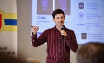 Чоловік у бордовій сорочці виступає перед аудиторією, на екрані за ним – презентація.