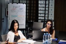 Дві жінки сидять за ноутбуком, слухаючи доповідача.