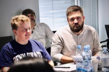 Двоє чоловіків сидять за столом і уважно слухають доповідача.
