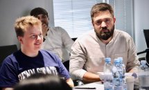 Двоє чоловіків сидять за столом і уважно слухають доповідача.