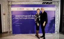 Двоє учасників заходу позують перед стендом із логотипами організаторів.