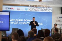 Спікер на сцені виступає перед аудиторією на тлі банера заходу.