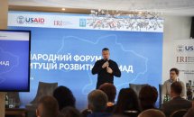 Спікер на сцені виступає перед аудиторією на тлі банера заходу.