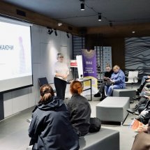 Учасники сидять у приміщенні з екраном та прапорами на задньому плані, слухаючи доповідача.