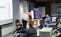 Учасники сидять у приміщенні з екраном та прапорами на задньому плані, слухаючи доповідача.