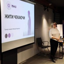 Жінка стоїть біля великого екрану з презентацією, на якому видно текст "Жити чекаючи".