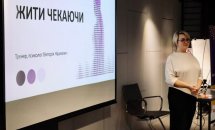 Жінка стоїть біля великого екрану з презентацією, на якому видно текст "Жити чекаючи".