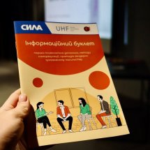 Людина тримає у руках буклет із заголовком "Інформаційний буклет", на обкладинці ілюстрації людей у діловій обстановці.