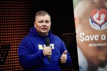 Чоловік у синьому худі говорить у мікрофон на тлі банера «Save a Life».кран із логотипом «Save a Life».м «Save a Life».