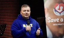 Чоловік у синьому худі говорить у мікрофон на тлі банера «Save a Life».