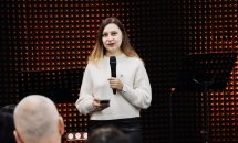 Жінка в білій кофтині виступає перед аудиторією з мікрофоном у руках.