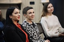  Аудиторія слухає виступ, дві жінки в передньому ряду посміхаються.