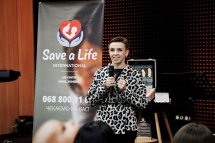 Жінка в чорно-білій сукні виступає на сцені, поруч екран із логотипом «Save a Life».