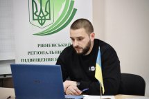 Молодий чоловік у чорному светрі працює за ноутбуком, поруч прапор України, на задньому фоні логотип організації.
