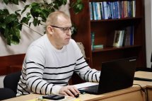 Чоловік у окулярах друкує на ноутбуці, навколо книжкові полиці та зелені рослини.