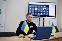 Учасник онлайн-зустрічі сидить перед ноутбуком із прапором України, на екрані позаду – учасники відеоконференції.