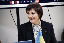 Усміхнена жінка в діловому костюмі сидить за столом, перед нею ноутбук, на екрані видно відеоконференцію.