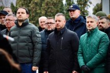 Група чоловіків у темному та зеленому одязі стоїть на відкритому повітрі, виглядають зосередженими.