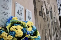Меморіальна стіна з портретами, біля якої покладено великий вінок із жовто-синіми квітами.