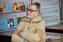 Жінка поважного віку у светрі уважно слухає доповідача, склавши руки на грудях. Позаду неї – стенд із навчальними матеріалами.