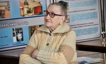 Жінка поважного віку у светрі сидить склавши руки на грудях. Позаду неї стенд