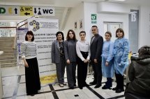 Група людей у медичних халатах і масках стоїть у приміщенні з табличками та вказівниками, ймовірно, у адміністративному закладі.