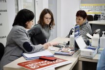 Дві жінки сидять за столом із документами та ноутбуком, одна з них пише, ймовірно, надаючи консультацію або реєструючи інформацію.