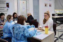 Дві особи в захисних медичних костюмах сидять за столом, беруть кров з пальця.