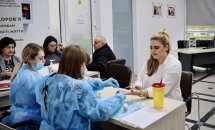 Дві особи в захисних медичних костюмах сидять за столом, беруть кров з пальця.
