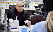 Людина похилого віку в пальто сидить за столом, спілкуючись із медичним працівником. 