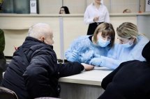 Дві особи, одна з них у темному пальті, сидять за столом із медичним персоналом у блакитних халатах, можливо, під час консультації чи оформлення документів.